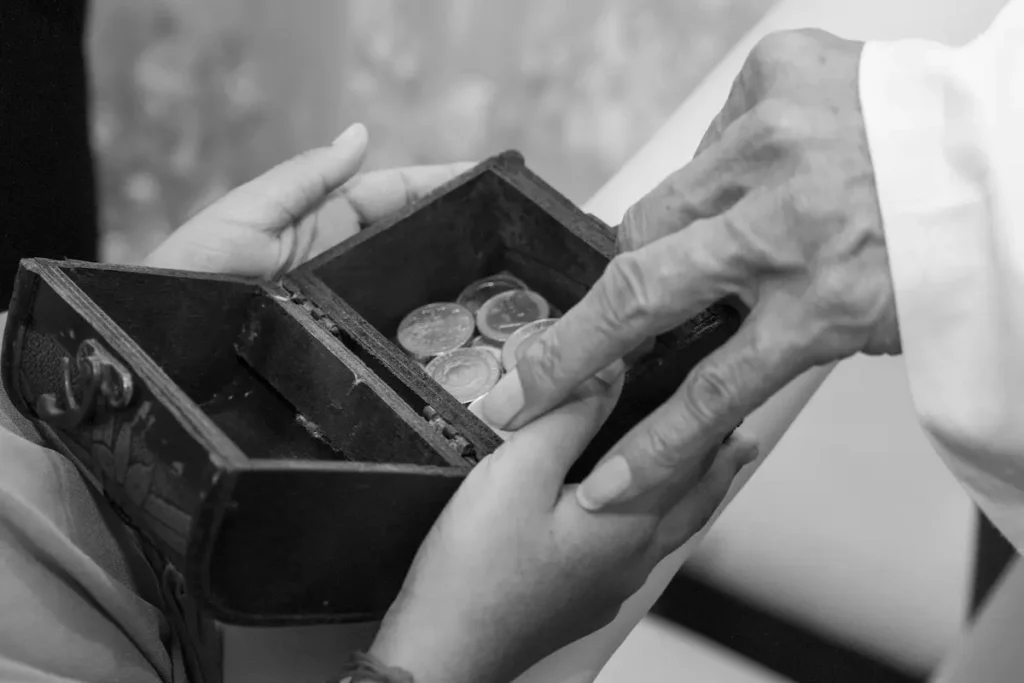 Black and white close-up of an older, wrinkled hand taking or giving coins from a small wooden box held by a younger person.